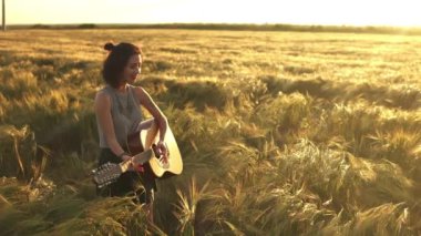 Güneşli yaz sahasında gitar çalan mutlu bir kadın. Beyaz bir bayan şarkı söylüyor ve eğleniyor. Ağır çekim. Küçük gitar. Müzik, doğa, yaşam tarzı konsepti. Müzisyen gitarist bayan gün batımını çalıyor..