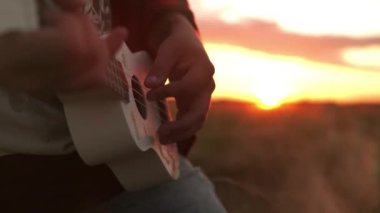 Genç hipster buğday tarlasında ukulele ile ritmik bir şekilde oynar. Güzel gün batımı. Kaygısız bir adam doğanın ve güneş ışığının tadını çıkarıyor. İnanılmaz renkli sarı günbatımında. Aşk özgürlüğü kavramı, yaşam tarzı