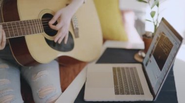 Genç Asyalı kadın evde gitar çalıyor. Müzisyen olmak isteyen biri dizüstü bilgisayarla müzik aleti çalmayı öğreniyor. Evde tek başına takıl. Güzel kız online ders izliyor ve müzik çalışıyor.