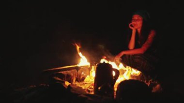 Genç turist kadın geceleri doğadaki şenlik ateşinin yanında dinleniyor. Yürüyüş yapan kızın parlak alevli yüzü. Turizm ve seyahat.