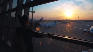 Havaalanı terminalinde büyük pencereye doğru yürüyen genç bir kadın. Turist yoğun havaalanının keyfini çıkarırken aynı zamanda muhteşem bir hava kararmasını bekliyor..