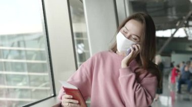 Havaalanı terminalinde maskeli yalnız bir kız karantinadan sonraki ilk uçuşundan son derece mutlu bir şekilde elinde biletle telefonda konuşuyor. Coronavirus önleme konsepti. Salgın sırasında seyahat.