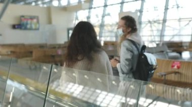 Tıbbi maskeli iki kadın havaalanı terminalinin lobisinde yürüyorlar, arka plan kavramı. Coronavirus sırasında uluslararası uçuşla gelen veya giden gezginler veya yolcular.