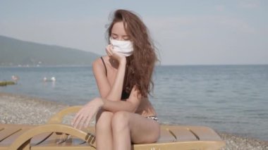 Bikini giyen üzgün genç kız virüs enfeksiyonunu önlemek için maske takmaya zorlandı. Mutsuz kadın, halk plajında tıbbi maske takmış ve kameraya bakıyor..