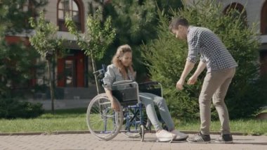 Genç adam tekerlekli sandalyedeki engelli kız arkadaşına ayakkabı bağcığı bağlıyor. Dokunaklı bir yardım ve destek anı. Birbirine aşık bir çift dışarıda yürüyor. Engelli yardımı kavramı. 6k küçültme 10 bit