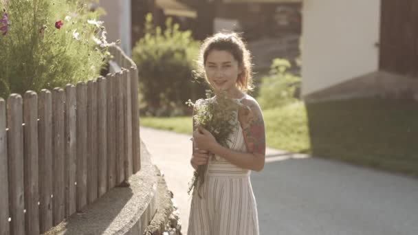 Portrét krásné usměvavé dívky s tetováním na silnici ve vesnici ve zpomaleném filmu. Žena s květinami v rukou tráví čas venku v létě nebo na jaře. Žena odpočívající v přírodě během dovolené.