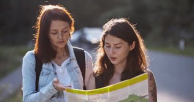 Kızlar bir harita ile yol boyunca yürüyorlar. Kadınlar gülümseyerek ziyaret etmek istedikleri yerler hakkında konuşuyorlar. Elinde bir rehberle seyahat eden iki güzel esmer. Yürüyüş turları, otostop çekmeler.