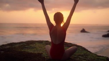 Yoga yapan genç bir kadının silueti. Okyanus kıyısında dinlenen kırmızı mayolu bir kız. Güzel esmer kadın, gün batımının önünde meditasyon yapıyor. Sağlık kavramı, rahatlama, yoga.