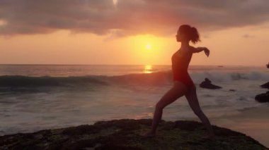 Mayo giymiş güzel bir kadın okyanus kıyısında yoga yapıyor. Dalgalar kuma vururken egzersiz yapan genç bir kızın silueti. Kadın güneş ışığı önünde meditasyon yapıyor. Sağlık kavramı, rahatlama, yoga.