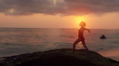 Sağlık kavramı, rahatlama, yoga. Mayo giymiş güzel bir kadın okyanus kıyısında yoga yapıyor. Dalgalar kuma vururken egzersiz yapan genç bir kızın silueti. Kadın gün ışığında meditasyon yapar..