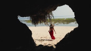 Karışık ırk bayanı tatilini tropik ada kayalık mağaralarında geçiriyor. Bali egzotik atmosferi. Güzel Asyalı kadın kırmızı elbiseyle tropikal beyaz kumsalda yürüyor. Güzellik, seyahat, moda konsepti.