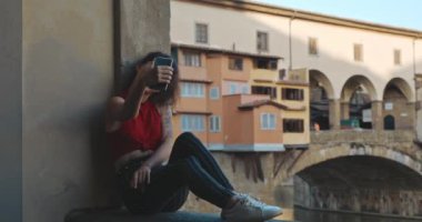 Yaz tatili sırasında Floransa 'ya seyahat eden genç bir kadının portresi. Kırmızı tişörtlü ve çizgili kot pantolonlu Asyalı kız Vecchio köprüsünde oturuyor ve akıllı telefonunda selfie çekiyor..