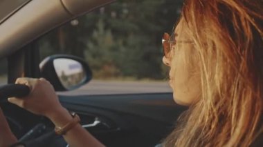Güzel kız seyahat ederken sakin müzik dinliyor ve dinleniyor. Gün batımında arabasını süren şık güneş gözlüğü takan sevimli sarışın yüzün yakın çekimi. Yaşam tarzı ve güzellik kavramı.
