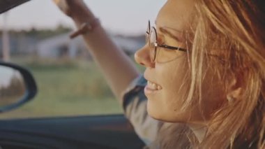 Güneş batarken arabasını süren şık güneş gözlüklü sevimli sarışın portresinin yakın çekimi. Güzel kız seyahat ederken sakin müzik dinliyor ve dinleniyor. Yaşam tarzı ve güzellik kavramı.