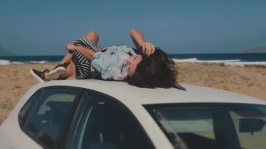 Magnifficent Sahili 'nde arabanın yakınında. Arabanın çatısında kaygısızca yatan güzel genç bir kadın. Sıcak yaz gününde saçlarına dokunarak hayatın tadını çıkaran bir kız. Seyahat, tatil, macera, gençlik, eğlence kavramı