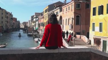 Köprünün kenarında oturan kırmızı kazaklı, çekici esmer kızın arkadan görünüşü süzülen gondollara ve kanal kenarında yürüyen turistlere bakıyor. İtalya 'da güneşin sıcacık ışınlarında dinlenmek.