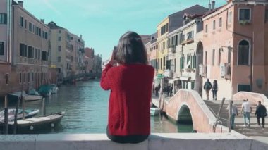 İtalya 'da güneşin sıcacık ışınlarında dinlenmek. Köprünün kenarında oturan kırmızı kazaklı çekici esmer kızın arkadan görünüşü süzülen gondollara ve kanal kenarında yürüyen turistlere bakıyor..