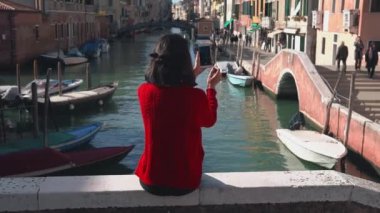 Kırmızı kazaklı güzel Asyalı kızın köprünün kenarından akıllı telefonuyla Venedik Kanalı 'nın fotoğrafını çekişi. Hoş bir kadın tatilini Venedik 'te geçiriyor..