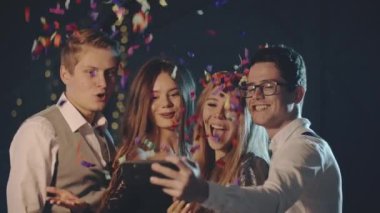 Gülümseyen çok ırklı arkadaşlar konfeti fırlatıp, akıllı telefondan veya cep telefonundan selfie çekiyorlar. Mutlu adamlar Noel partisinde modern teknolojiyi kullanarak eğleniyor ve fotoğraf çekiyorlar..
