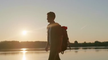 Gün doğumunda nehrin kenarında büyük bir sırt çantasıyla yürüyüş yapan genç bir adamın silueti. Yolculuğun tadını çıkaran bir adam. Kendine karşı zafer kazanıyorsun. Yürüyüş kavramı, sağlıklı yaşam tarzı, seyahat. Tek başına etkinlik