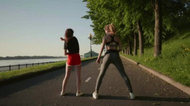 Güzel kızlar nehir kenarında koşmadan önce kaslarını esnetiyorlar. Genç seksi kadınlar birlikte uzun mesafe koşmaya başlarlar. Eğitimden önce ısın. Yürüyüş kavramı, zayıf uyum, egzersiz..