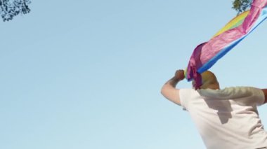 Mavi gökyüzünün arka planında geçit töreni yaparken elinde gökkuşağı eşcinseli bayrağı tutan eşcinsel adam. Kalp gözlüğü takan mutlu adam haklarını gösteriyor. LGBTQI, Gurur Etkinliği, LGBT Gurur Ayı, Eşcinsel Onur Sembolü