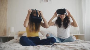 Sanal gerçeklik kulaklığı takmış iki kadın yatakta oturmuş, video oyunları oynuyor ve birlikte eğleniyorlar. Evde sanal gerçeklik kasklı kızlar var. Üç boyutlu teknoloji. Sanal gerçeklik, oyun, arkadaşlık kavramı.