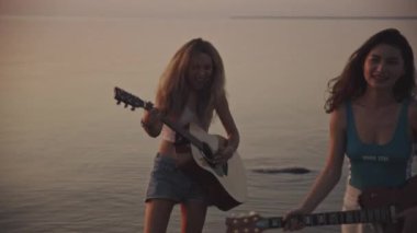 Gün batımında deniz kıyısında gitar çalarak eğlenen uluslararası bir çift kız. Güzel Asyalı ve sarışın kadın gülümsüyor ve müzik yapıyor. Arkadaşlarla tatil..