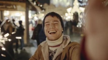 Eski Avrupa şehrinde Noel arifesinde muhteşem sokak fuarında gülümseyen ve akıllı telefonuyla selfie çeken komik genç adam. Arka planda sıradan insanlar yürüyor ve hediyelik eşya alıyor..
