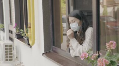 Koruyucu maskeli üzgün Asyalı kadın pencereden dışarı bakıyor. Kendini Karantina 'da Evde Kalmak. Covid-19, koronavirüs pandemik konsepti. 6k küçültme 10 bit
