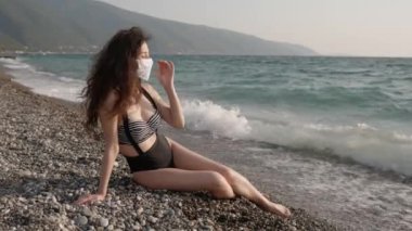 Halk plajında koruyucu cerrahi maske takan ve hayatın tadını çıkaran güzel bir kadının portresi. Koronavirüse karşı önlem kavramı. Salgın sırasında sağlık ve güvenlik, N1H1