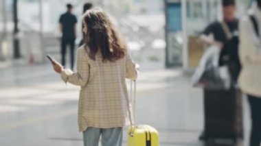 Havaalanı terminali boyunca elinde akıllı telefonla hareket eden bir iş kadınının görüntüsü. Genç bir kadın bavuluyla yürüyor ve haberleri kontrol ediyor ya da telefonda sohbet ediyor. Seyahat kavramı, teknoloji.