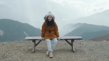 Soğuk havada bulutlu dağlarda yalnız bankta oturan mutlu Asyalı kadın turist. Dağ manzarasının dumanlı arka planında genç bir yürüyüşçü. Etkin yaşam biçimi kavramı