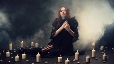 Kızıl saçlı cadı duman içinde oturmuş şeytanları çağırmak için enerjik el hareketleri yapıyor. Şeytan 'ı çağırmak için büyülü bir ayin. Cosplay, cadılar bayramı kostümü. Doğaüstü, kötü, karanlık, gotik kavramı.