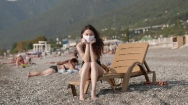 Halka açık plajda koruyucu maske takan, güverte sandalyesinde oturan mutsuz, üzgün, melez bir ırkın portresi. Koronavirüse karşı önlem kavramı. Salgın sırasında sağlık ve güvenlik hayatı, N1H1