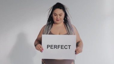 Doğal mükemmel değil. İç çamaşırlı mutlu şişman kadın elinde pozitif vücut posteri tutuyor. Çeşitli gerçek güzellik, çeşitlilik, kusurlu tolerans konsepti. Cesur kız kameraya gülümsüyor..