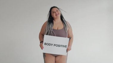 Çeşitli güzellik, çeşitlilik hoşgörü konsepti. İç çamaşırlı mutlu şişman kadın elinde pozitif vücut posteri tutuyor. Tatlandırıcıdaki şişman kız kameraya gülümsüyor..