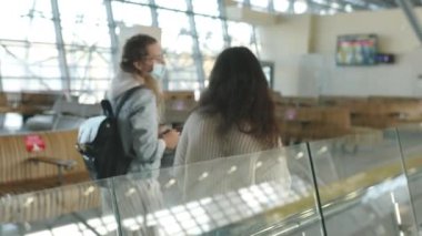 Tıbbi maskeli iki kadın havaalanı terminalinin lobisinde yürüyorlar, arka plan kavramı. Coronavirus sırasında uluslararası uçuşla gelen veya giden gezginler veya yolcular.