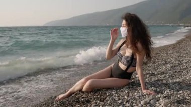 Halk plajında koruyucu cerrahi maske takan ve hayatın tadını çıkaran güzel bir kadının portresi. Koronavirüse karşı önlem kavramı. Salgın sırasında sağlık ve güvenlik, N1H1