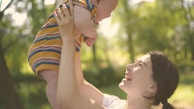 Mutlu anne, oynamak ve dünyayı gezdirmek için ellerini havaya kaldırıp çocuğu kaldırıyor. Çocuk ve ebeveyn birlikte parkta yürürken gülümsüyor. Anne ve bebek birlikte oynuyorlar..