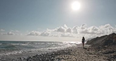 Güzel kız yavaşça sahilde hareket ediyor. Kayalık sahilde siyah mayo giymiş tatlı bir esmer. Resimli manzaranın arka planında büyüleyici bir kadın silueti. Güzellik ve film kavramı.