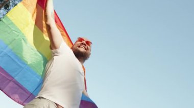 Mavi gökyüzünün arka planında geçit töreni yaparken elinde gökkuşağı gay bayrağı tutan eşcinsel bir adam. Kalp gözlüğü takan mutlu adam haklarını gösteriyor. LGBTQI, Gurur Etkinliği, LGBT Gurur Ayı, Eşcinsel Onur Sembolü