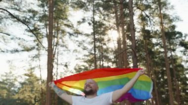 Orman parkında gökkuşağı gay bayrağıyla koşan eşcinsel bir adam. Mutlu adam haklarını gösteriyor. Utanmaz ibne cinsel tercihini gösteriyor. LGBTQI, Gurur Etkinliği, LGBT Gurur Ayı, Eşcinsel Onur Sembolü