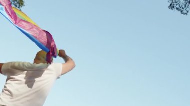 Mavi gökyüzünün arka planında geçit töreni yaparken elinde gökkuşağı eşcinseli bayrağı tutan eşcinsel adam. Kalp gözlüğü takan mutlu adam haklarını gösteriyor. LGBTQI, Gurur Etkinliği, LGBT Gurur Ayı, Eşcinsel Onur Sembolü