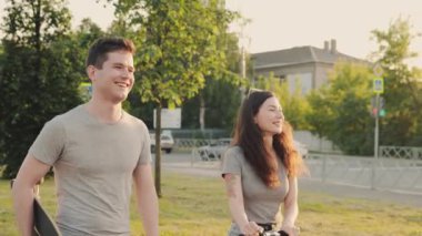 Arkadaşlar yürürken birbirleriyle iletişim kurarlar. Güzel Asyalı kız ve yakışıklı adam turuncu gün batımının önünde mimari binadan ya da parktan ellerinde kaykayla geçiyorlar..