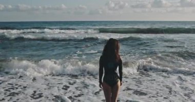 Siyah mayo giymiş seksi bir kız silueti. Genç, güzel kadın poposu denize doğru yürüyor. Doğal manzaranın arka planında narin kız kalçaları..