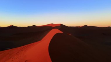 Afrika çölündeki turuncu kum tepeciklerinin üzerinden insansız hava aracı atışı. Açık mavi alacakaranlık gökyüzünün altındaki kumdan tepenin muhteşem hava manzarası. İnsansız hava aracı tarafından yakalanan çorak yabanın renkli kontrastı.