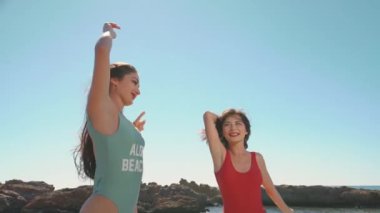 Muhteşem deniz sahillerinde poz veren iki güzel genç kız. Sıcak yaz melteminin tadını çıkaran kadınlar, mavi gökyüzünün altında kumlu sahilde zıplayan ve el sallayan kadınlar. Tatil, seyahat, gençlik kavramı.