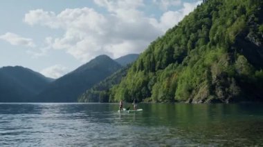 Kürek tahtası maceraları. Gün batımında dağ eteğinde yemek güvertesinde sörf yapan iki kadın. Yaz tatili boyunca bayan arkadaşlar. Resimli bir manzara. Spor ve turizm konsepti.