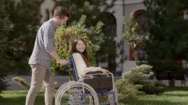 Genç adam sakat kız arkadaşını tekerlekli sandalyede sürerken konuşuyor ve gülüyor. Felç olmuş, hayatı dolu dolu yaşayan Asyalı engelli bir kız. Birbirine aşık bir çift. 6k aşağı ölçek 10 bit renk.